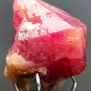 May include: A vibrant pink, faceted, rough-cut gemstone with a slightly translucent appearance. The gemstone is held in a metal clamp.