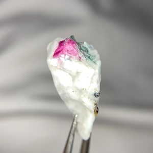 May include: A small, rough-cut gemstone with a white base, featuring vibrant pink and green inclusions. The stone is held by silver tweezers, showcasing its natural, unpolished texture. Ideal for jewelry making or collecting.