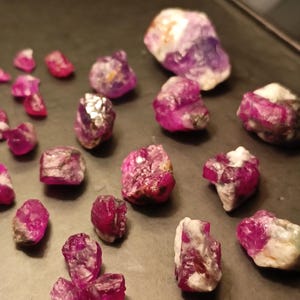Puede incluir: Una colección de piedras preciosas de rubí en bruto, que muestran un vibrante tono rosa-rojizo. Las piedras en bruto varían en tamaño y forma, con algunas que muestran inclusiones blancas y grises. Las gemas están dispuestas sobre una superficie oscura.