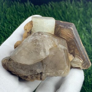 May include: A collection of raw, natural crystals and minerals. The image shows a cluster of translucent quartz crystals, a light-colored rectangular crystal, and a brown, angular mineral formation. The crystals are set against a backdrop of green grass.