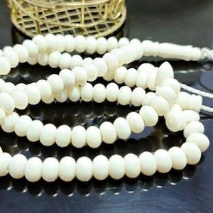 May include: A white prayer bead necklace with oval beads. The beads are strung together with a white cord, forming multiple loops. The necklace is resting on a reflective black surface, with a gold-coloured decorative object in the background.