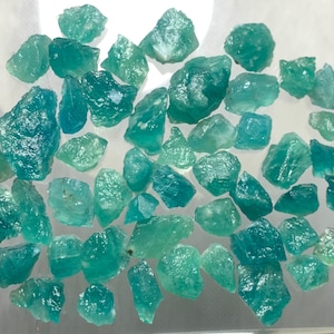 Op de afbeelding: Een verzameling van talrijke ruwe, ongepolijste edelsteenfragmenten in verschillende tinten blauwgroen, turkoois en zeegroen. Deze kleine, grillige mineraalstukjes vertonen een doorschijnende kwaliteit met onregelmatige, kristallijne texturen.