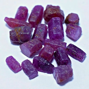 May include: A collection of raw, purple ruby gemstones. The rough stones are cut into rectangular shapes and have a natural, unpolished finish.
