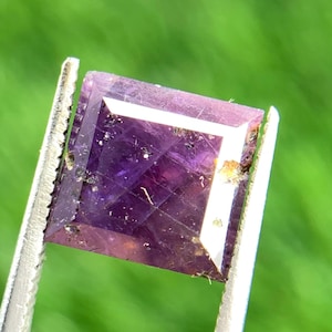 Könnte beinhalten: Ein quadratisch geschliffener, facettierter Amethyst-Edelstein, der von einer Pinzette gehalten wird. Der Edelstein hat eine tiefviolette Farbe mit einem weißen Rand und einer reflektierenden Oberfläche. Der Hintergrund ist ein verschwommenes Grün.