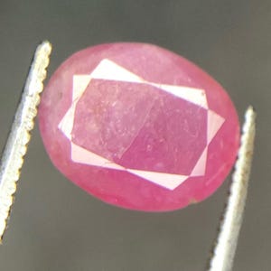 Op de afbeelding: Een gefacetteerde, ovaalvormige robijn edelsteen, vastgehouden door een pincet. De robijn heeft een levendige roze-rode kleur met een geometrisch, wit gefacetteerd ontwerp in het midden. De pincet is zilverkleurig en de achtergrond is een vage grijstint.