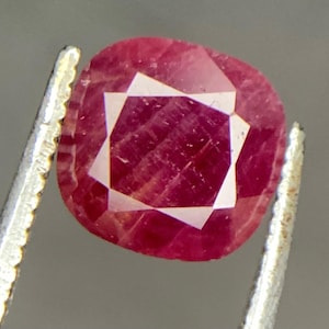 May include: A faceted, cushion-cut ruby gemstone held by silver tweezers. The ruby is a deep, saturated red with a square, white, star-like pattern in the center. The gemstone's facets reflect light, creating a sparkling effect.