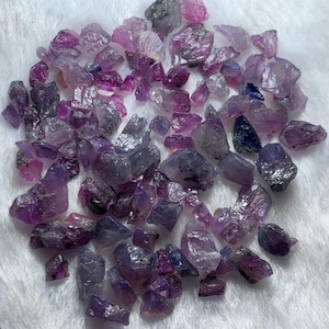 May include: A collection of raw, unpolished sapphire gemstones in various shades of purple, violet, and deep blue. These small, jagged mineral fragments exhibit natural crystalline textures, translucent to opaque clarity, and irregular geometric shapes.