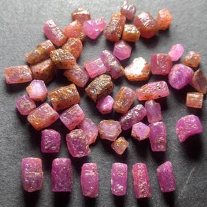 May include: A collection of rough-cut ruby gemstones in various shapes and sizes. The gemstones display a range of pink and reddish-brown hues, with a slightly textured surface. The stones are scattered across a dark gray surface.