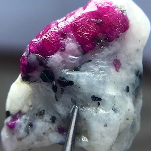 May include: A close-up of a pink ruby crystal embedded in a white matrix. The crystal has a rough, natural texture and is surrounded by black specks.