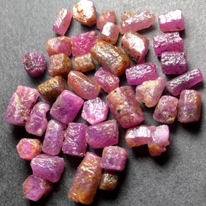 Puede incluir: Una colección de piedras preciosas de rubí en bruto de varias formas y tamaños. Las piedras muestran una gama de tonos rosados y rojo-púrpura, algunos con un color marrón dorado. La textura rugosa y las formas naturales de los rubíes son visibles.
