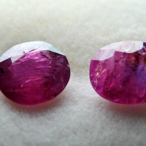 May include: Two round, faceted, pink gemstones with a slight purple hue. The gemstones are set on a white background.