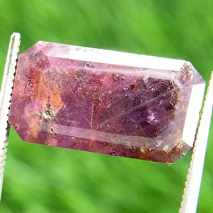 May include: A faceted, rectangular gemstone with a deep pink hue, held by silver tweezers against a blurred green background. The gem has a transparent quality with internal inclusions and a polished surface, showcasing its natural beauty.