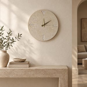 Könnte beinhalten: Eine runde Wanduhr aus beigefarbenem, strukturiertem Stein mit gesprenkelter Oberfläche. Das Zifferblatt hat eine dreieckige Markierung auf der Zwölf-Uhr-Position, rechteckige Markierungen bei drei, sechs und neun sowie zwei dünne goldfarbene Zeiger.