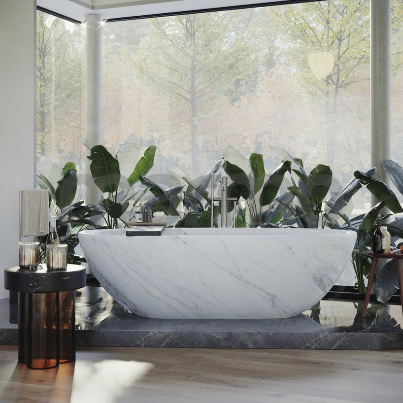 Op de afbeelding: Een wit marmeren bad met een modern ontwerp staat in een badkamer met grote ramen en weelderige groene planten. Het bad is vrijstaand en heeft een strakke, minimalistische uitstraling.