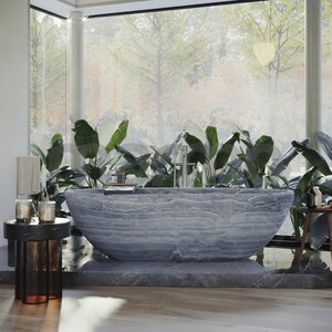 Op de afbeelding: Een groot, grijs stenen bad staat in een moderne badkamer met grote ramen en weelderige groene planten. Het bad heeft een glad, getextureerd oppervlak en is omgeven door een grijze tegelvloer.