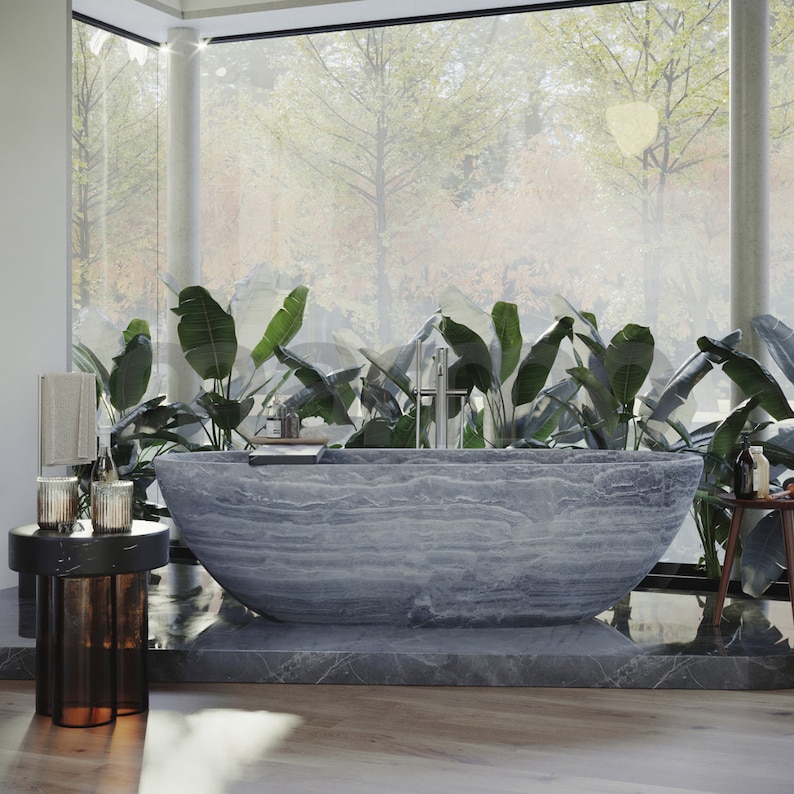 Op de afbeelding: Een groot, grijs stenen bad staat in een moderne badkamer met grote ramen en weelderige groene planten. Het bad heeft een glad, getextureerd oppervlak en is omgeven door een grijze tegelvloer.