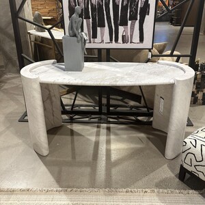 Può includere: Tavolo consolle in marmo bianco con piano curvo e bordi arrotondati. Il tavolo ha un design moderno ed è sostenuto da due gambe spesse e arrotondate.