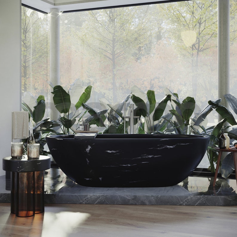 Op de afbeelding: Een zwart marmeren bad met een strak, modern ontwerp staat in een badkamer met grote ramen en weelderige groene planten. Het bad is geplaatst op een verhoogd platform met een grijs marmeren oppervlak.