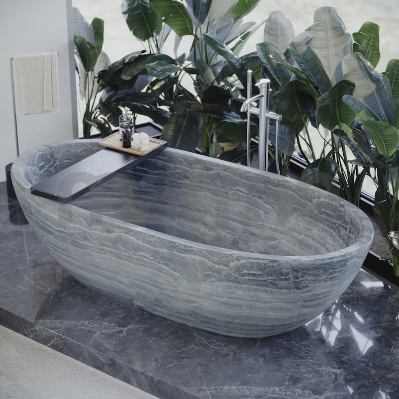 Op de afbeelding: Een groot, ovaalvormig bad van grijs steen met een houten plank aan de zijkant. Het bad staat in een badkamer met een raam erachter, waar weelderige groene planten te zien zijn.