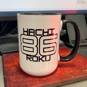 May include: White ceramic mug with a black rim and handle. The mug has a black graphic that says "Hachi 86 Roku" on the front.