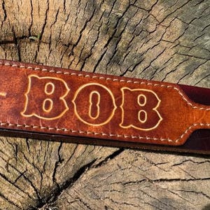 May include: A brown leather strap featuring white stitched edges and the name BOB embossed in a bold, stylized font. The leather has a smooth, polished finish with visible grain texture and a slightly darker, reddish-brown tone along the edges.
