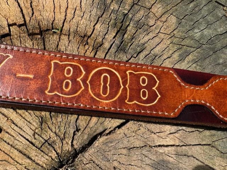 May include: A brown leather strap featuring white stitched edges and the name BOB embossed in a bold, stylized font. The leather has a smooth, polished finish with visible grain texture and a slightly darker, reddish-brown tone along the edges.
