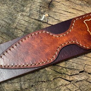 May include: A dark brown leather belt featuring a lighter tan leather overlay with a pointed, elongated shape. The overlay is secured with visible light-colored stitching along its edges. A portion of a letter is visible on the tan leather section.
