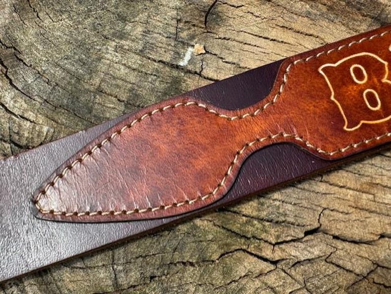 May include: A dark brown leather belt featuring a lighter tan leather overlay with a pointed, elongated shape. The overlay is secured with visible light-colored stitching along its edges. A portion of a letter is visible on the tan leather section.