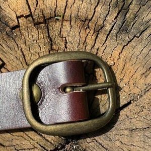 May include: A dark brown leather belt featuring a smooth, matte finish and a rounded, antique brass-toned metal buckle. The belt strap is secured to the buckle frame with a single matching brass-toned metal rivet, creating a clean, functional design.