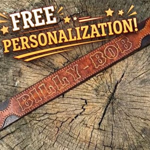 May include: A brown leather belt featuring a pointed end design and the name BILLY-BOB embossed in a western-style font. The text FREE PERSONALIZATION! is displayed in yellow letters above the belt, which is positioned on a textured wood surface.