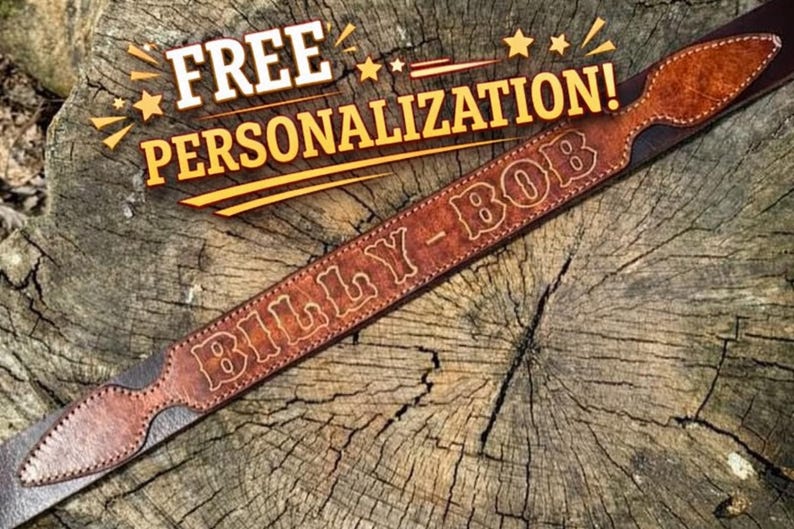 May include: A brown leather belt featuring a pointed end design and the name BILLY-BOB embossed in a western-style font. The text FREE PERSONALIZATION! is displayed in yellow letters above the belt, which is positioned on a textured wood surface.