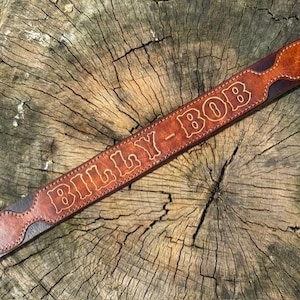May include: A brown leather belt featuring a decorative overlay with a pointed end. The leather is embossed with the text BILLY-BOB in a bold, outlined font. The belt is constructed with visible stitching along the edges of the overlay.