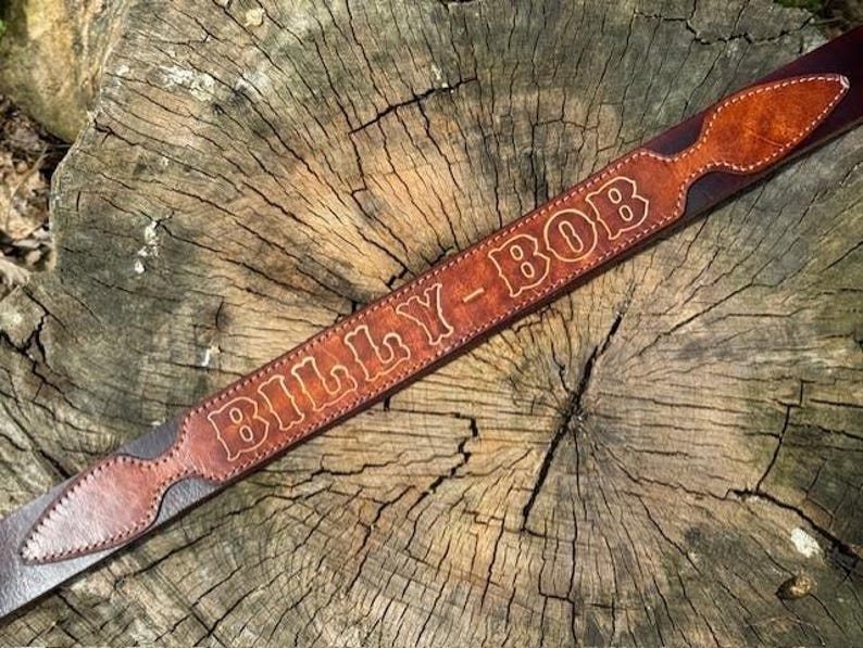 May include: A brown leather belt featuring a decorative overlay with a pointed end. The leather is embossed with the text BILLY-BOB in a bold, outlined font. The belt is constructed with visible stitching along the edges of the overlay.