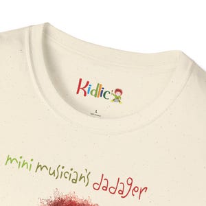 May include: Cream-coloured t-shirt with the text "Kidlic" in colourful letters at the neckline. Below, the words "mini musician's dadager" are written in green and red. The shirt is made of 100% cotton.