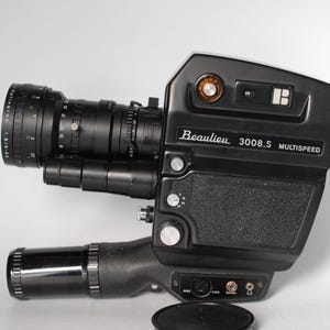 Könnte beinhalten: Schwarze Beaulieu 3008.S Multispeed Filmkamera mit großem Objektiv. Die Kamera hat einen strukturierten Griff und mehrere Drehknöpfe und Tasten. Das Objektiv ist schwarz mit silbernen Markierungen. Unter der Kamera liegt ein schwarzer Objektivdeckel.