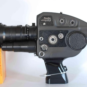 Könnte beinhalten: Eine graue Beaulieu 4008 ZM II Filmkamera mit schwarzem Objektiv und Griff, platziert neben einer gelb-roten Schachtel Kodachrome 40 Film. Die Kamera verfügt über mehrere runde Einstellräder, einen seitlichen Objektivdeckel und eine schwarze Augenmuschel.