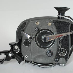 May include: A vintage gray and black movie camera with a handle, viewfinder, and various dials. The camera has a silver crank and a textured surface. The handle is black and has a curved design. The camera is on a white background.