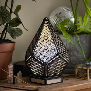 Puede incluir: Una lámpara geométrica negra con un diseño estampado en blanco y negro. La lámpara está encendida y proyecta un brillo cálido. La lámpara está sobre una mesa de madera con plantas y otros elementos decorativos.