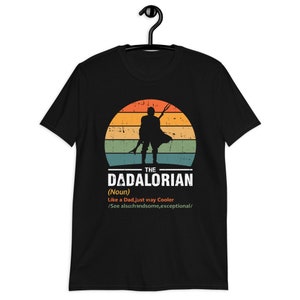 May include: Black t-shirt featuring a silhouette of a figure against a sunset backdrop. Text on the shirt reads "THE DADALORIAN (Noun) Like a Dad, just way Cooler /See also: handsome, exceptional/". The shirt is displayed on a black hanger.