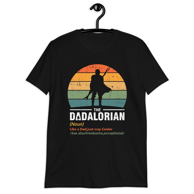 May include: Black t-shirt featuring a silhouette of a figure against a sunset backdrop. Text on the shirt reads "THE DADALORIAN (Noun) Like a Dad, just way Cooler /See also: handsome, exceptional/". The shirt is displayed on a black hanger.