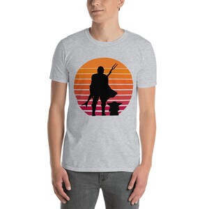 May include: A grey t-shirt with a silhouette of a character holding a weapon and a small creature standing next to them. The silhouette is in front of a sunset with orange, red, and yellow stripes.
