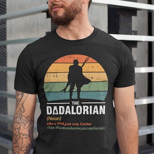 May include: Dark grey t-shirt with a retro sunset design and a silhouette of a figure holding a weapon. The text reads "THE DADALORIAN (Noun) Like a Dad, just way Cooler /See also: handsome, exceptional/."