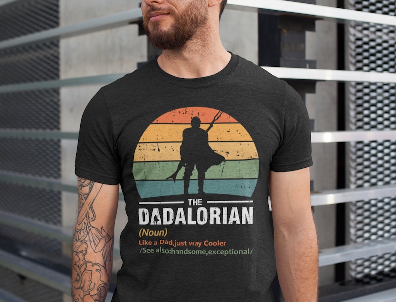 May include: Dark grey t-shirt with a retro sunset design and a silhouette of a figure holding a weapon. The text reads "THE DADALORIAN (Noun) Like a Dad, just way Cooler /See also: handsome, exceptional/."