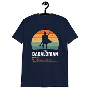 May include: Dark blue t-shirt with a retro sunset design and a silhouette of a figure holding a weapon. The text reads "THE DADALORIAN" with a definition below. A unique graphic tee for fans of the series.