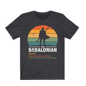 May include: Dark grey t-shirt with a retro sunset design and a silhouette of a figure with a weapon. The text reads "THE DADALORIAN (Noun) Like a Dad, just way Cooler /See also: handsome, exceptional/."