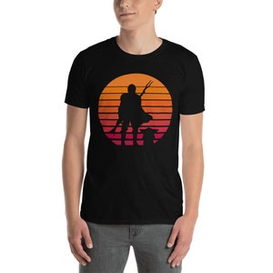 May include: A black t-shirt with a retro sunset graphic featuring a silhouette of a Mandalorian with a spear and Grogu. The graphic is in shades of orange, red, and pink.