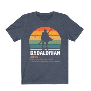 May include: Dark blue t-shirt with a graphic of a figure holding a weapon against a sunset. The text reads "THE DADALORIAN (Noun) Like a Dad, just way Cooler /See also: handsome, exceptional/."
