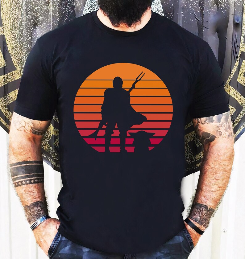 May include: A black t-shirt with a retro sunset graphic featuring a silhouette of a Mandalorian character holding a spear and a small creature.