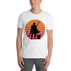 May include: White t-shirt with a silhouette of a character holding a weapon and a small creature standing in front of them. The silhouette is in front of a sunset with orange, red, and yellow stripes.
