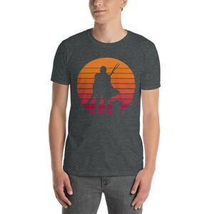 May include: A dark heather grey t-shirt with a retro sunset graphic featuring a silhouette of a Mandalorian character with a spear and a small creature.